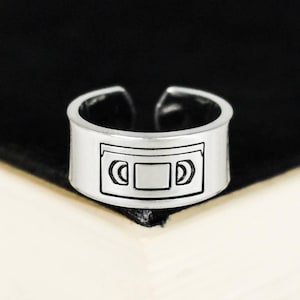Peut inclure: Bague réglable argentée avec un motif noir et blanc de cassette VHS. La bague a une bande épaisse et un design ouvert. Un accessoire sur le thème rétro.