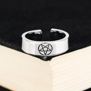 May include: A silver ring with a pentagram design. The ring is on a black surface.