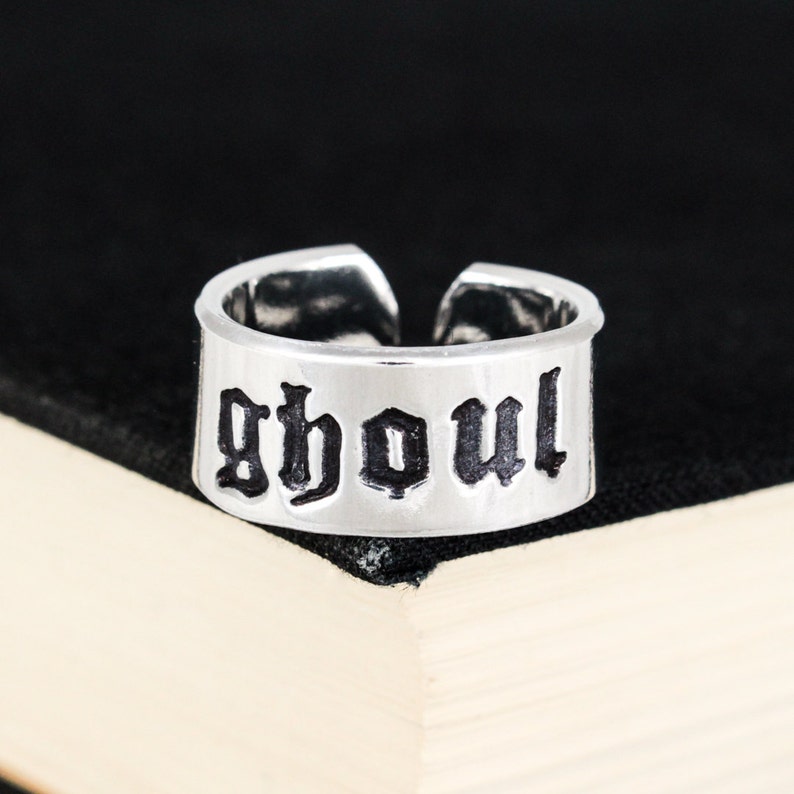 May include: A silver ring with the word "ghoul" written in black gothic lettering.