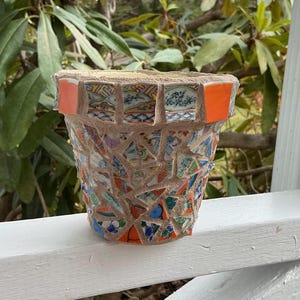 May include: A handcrafted mosaic flower pot. The pot is made from broken ceramic pieces in various colors, including blue, orange, and white. The rim features orange tiles and decorative ceramic pieces. The pot is sitting on a white wooden surface.