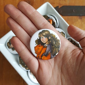 May include: A round button with a white background featuring a cartoon illustration of a brown-haired woman with a red cat in her arms. The woman is wearing a blue and white uniform and has a friendly expression. The cat is orange and white and has a grumpy expression.