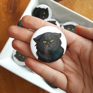 Könnte beinhalten: Ein weißer Knopf mit einer schwarzen Katze mit gelben Augen. Der Knopf wird in einer Hand gehalten.