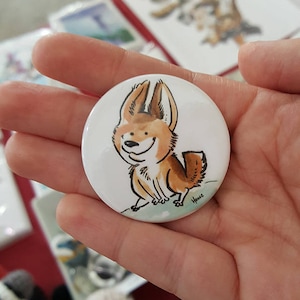 May include: A white button featuring a cartoon illustration of a fox with brown fur and a white belly. The fox is smiling and has large ears. The button is 6.35 cm in diameter.