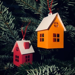Peut inclure: Deux décorations de Noël en papier en forme de maisons. La plus grande maison est orange avec des bordures blanches et un toit blanc. La plus petite maison est rouge avec des bordures blanches et un toit blanc. Les deux maisons ont des fenêtres et des portes.