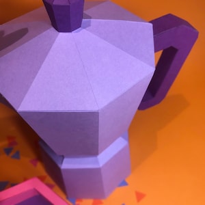 May include: A purple papercraft model of a coffee pot with a detailed lid and handle. The model is sitting on a bright orange surface with colorful confetti.