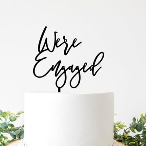 Peut inclure: Un cake topper noir avec les mots "We're Engaged" en écriture cursive. Le cake topper est placé sur un gâteau blanc glacé, décoré de verdure.