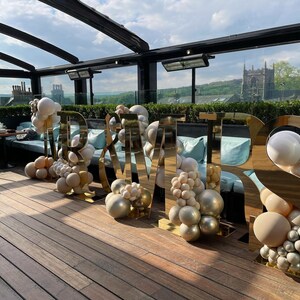 May include: Gold "Mr & Mrs" letters with balloon decorations on a wooden deck. The letters are reflective and the balloons are white, beige, and gold.