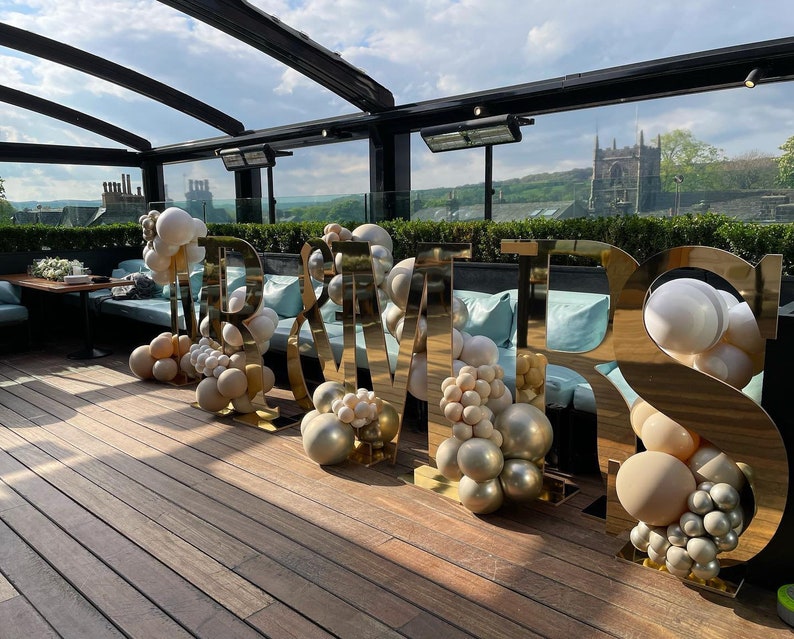 May include: Gold "Mr & Mrs" letters with balloon decorations on a wooden deck. The letters are reflective and the balloons are white, beige, and gold.