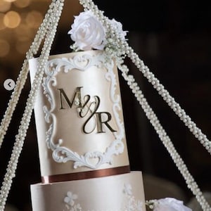 May include: A two-tiered wedding cake decorated with white pearl beading and a white floral accent. The top tier features a white and gold ornate frame with the initials "M & R" in gold.