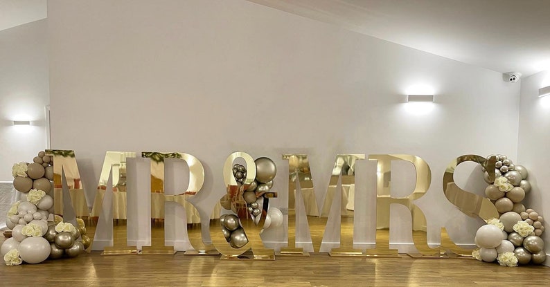 May include: Gold mirrored letters spelling "MR & MRS" with white and beige balloons on either side.