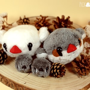 Pode incluir: Dois brinquedos de pássaros de pelúcia, um branco com um bico vermelho e olhos pretos, e outro cinza com um bico vermelho e olhos pretos. Os brinquedos estão sentados em uma superfície de madeira com pinhas.