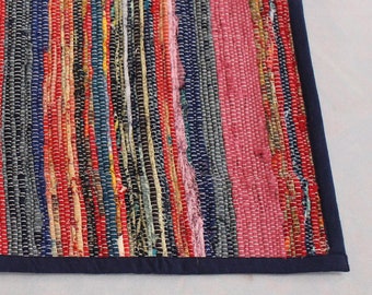 May include: Multicolored woven rug with a navy blue border. The rug has a striped pattern with various colors including red, pink, blue, green, and yellow.