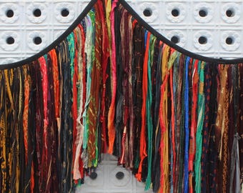 May include: A colorful, handmade textile wall hanging. The piece features a black curved top with numerous vertical strips of fabric in various colors, including red, orange, green, and blue. The wall hanging is suspended from wooden pegs.