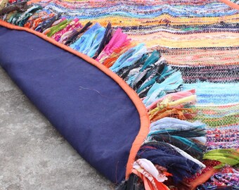 May include: A round, multi-colored rug with a fringe border. The rug is made of woven fabric and has a variety of colors, including blue, green, red, orange, and yellow. The rug is folded in half and the underside is a solid navy blue.