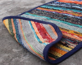 May include: A colorful, striped rug with a navy blue border. The rug is made of recycled materials and has a hand-woven texture.