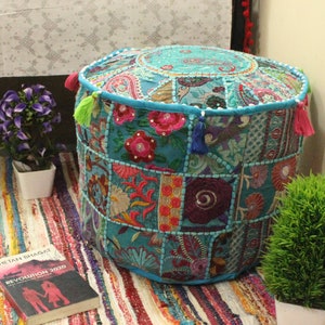 May include: A turquoise blue round pouf ottoman with colorful patchwork fabric and embroidered details. The pouf is sitting on a multi-colored striped rug.
