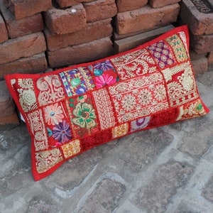 May include: A red patchwork lumbar cushion with gold embroidery and colourful floral patterns. The cushion is made of various fabrics and textures, creating a unique and bohemian style.