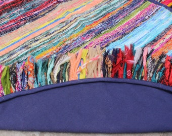 May include: A round, multi-colored rug with a fringe border. The rug is made of woven fabric and has a navy blue backing.