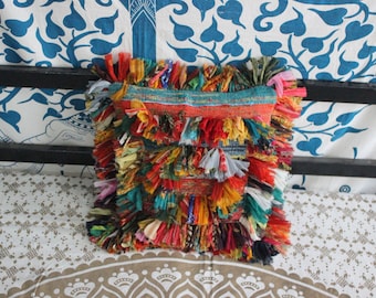 May include: A colorful, square pillow with a fringed edge. The pillow is made of various fabrics in shades of red, yellow, orange, and blue. The top edge has a band of orange and teal fabric.
