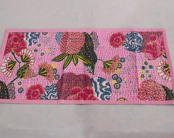 May include: Pink table runner with a floral pattern featuring pink, green, and blue flowers. The runner has a pink border.