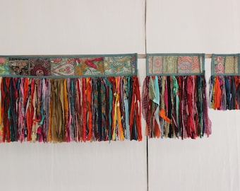 May include: A colorful fabric wall hanging with a patchwork design and long fringe. The hanging is made of various fabrics in shades of red, orange, yellow, green, blue, purple, and brown. The fringe is made of thin strips of fabric in the same colors.