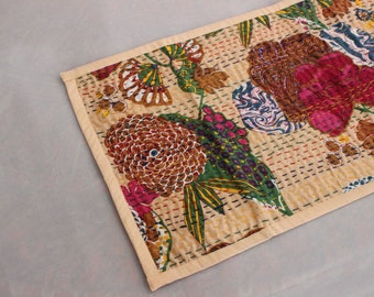 May include: A beige table runner with a floral pattern. The pattern features large pink, brown, and green flowers with intricate details. The runner has a cream-colored border.