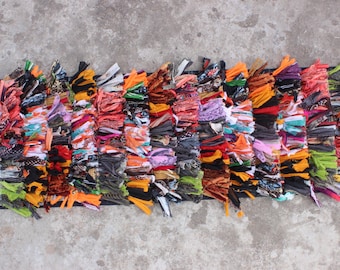 May include: A colorful, multi-colored, hand-woven rug with a fringe border. The rug is made of various fabrics and textures, including cotton, wool, and silk. The rug is approximately 3 feet long and 1 foot wide.