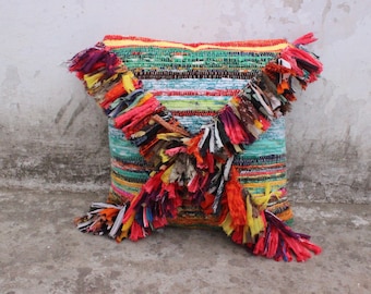 May include: A colorful throw pillow with a woven design and fringe trim. The pillow is made of a variety of fabrics and colors, including red, green, blue, yellow, and white.