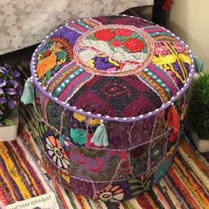 Puede incluir: Un puf redondo de color morado y multicolor con diseños florales bordados y borlas. El puf está hecho de retazos de tela cosidos juntos en un estilo patchwork.