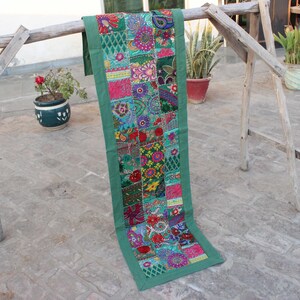 Puede incluir: Un camino de mesa verde con un diseño de patchwork que presenta bordados florales coloridos. El camino de mesa tiene un borde verde y está hecho de tela.
