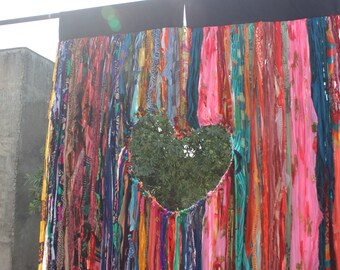 May include: A colorful fabric curtain with a heart-shaped opening revealing a green tree in the background. The curtain is made of many different colored strips of fabric, creating a textured and vibrant look.