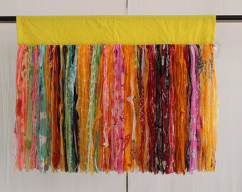 May include: A yellow fabric valance with a long fringe of colorful fabric strips. The fringe is made of various colors and patterns, including pink, red, orange, yellow, green, blue, and purple.