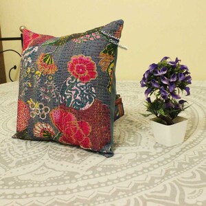 May include: A gray cotton pillow cover with a floral pattern in shades of pink, green, and yellow. The pillow cover is quilted and has a frayed edge. The pillow is sitting on a white bedspread with a mandala pattern.
