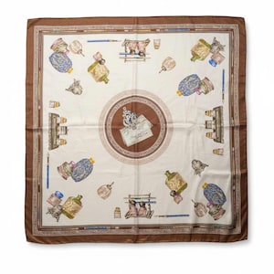 Puede incluir: Pañuelo cuadrado de seda con fondo crema y borde marrón. El pañuelo presenta un patrón repetido de tinteros, plumas e instrumentos de escritura en varios colores. Un diseño circular en el centro muestra una carta y elementos decorativos.