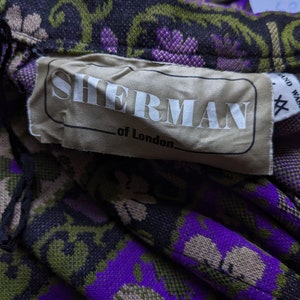 Puede incluir: Primer plano de una etiqueta de tela con el texto "Sherman of London" en letras blancas sobre un fondo beige. La etiqueta est&aacute; unida a una tela estampada en morado, verde y negro.