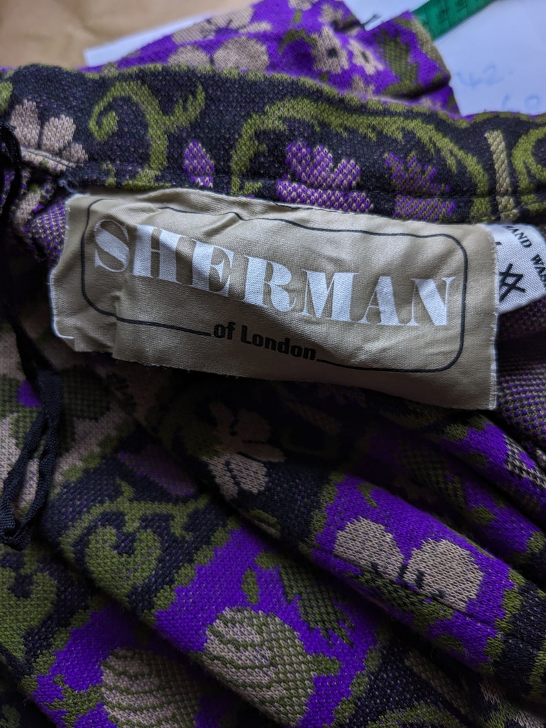 Puede incluir: Primer plano de una etiqueta de tela con el texto "Sherman of London" en letras blancas sobre un fondo beige. La etiqueta est&aacute; unida a una tela estampada en morado, verde y negro.