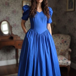 May include: A vibrant royal blue midi dress featuring short puff sleeves, a V-neckline, and a fitted bodice that transitions into a full, pleated skirt. The dress includes a distinct V-shaped waistline seam and side pockets for functional utility.