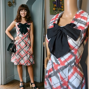 May include: A vintage-style dress with a red, white, and grey plaid design. It has a large black bow at the neckline. The dress is sleeveless and falls to the knee, with a fitted bodice.