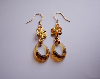 Könnte beinhalten: Ein Paar goldfarbene Hängeohrringe. Jeder Ohrring hat oben ein goldfarbenes Blumendesign, das mit einem klaren, oval geformten Anhänger mit braunen Sprenkeln verbunden ist. Die Ohrringe hängen an goldfarbenen Ohrhaken.