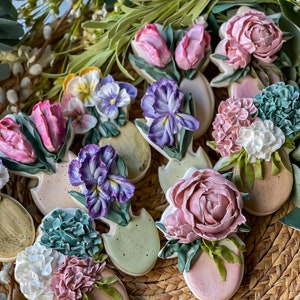 May include: A collection of decorated sugar cookies shaped like flower pots with various spring flowers, including tulips, pansies, irises, peonies, and hydrangeas. The cookies are arranged on a woven basket.