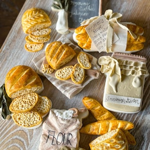 May include: A collection of miniature bread and baking supplies, including a bag of flour, a sourdough starter, a loaf of bread, and a baguette. The items are made of ceramic and painted in realistic colors.