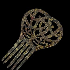 May include: A decorative hair comb with a heart-shaped top and a series of tines. The comb features a swirling, patterned design in shades of gold, brown, and green against a black background. The comb is a hair accessory.