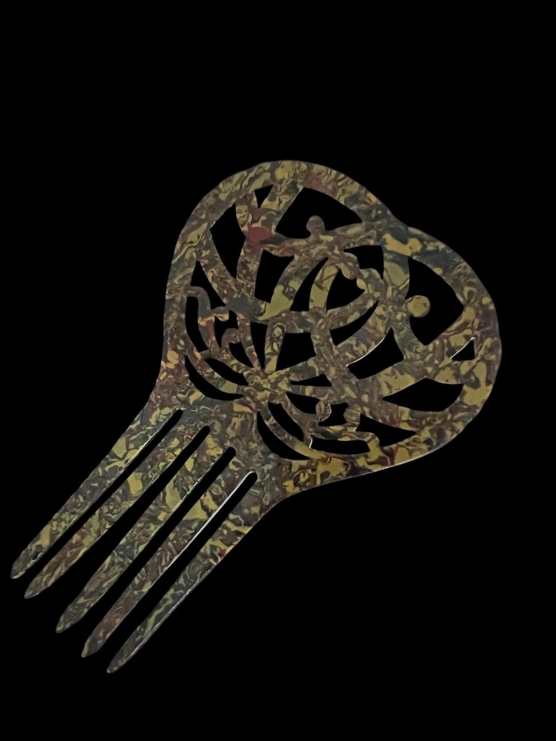 May include: A decorative hair comb with a heart-shaped top and a series of tines. The comb features a swirling, patterned design in shades of gold, brown, and green against a black background. The comb is a hair accessory.