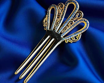 May include: A decorative hair comb featuring a fan-shaped top adorned with small, circular blue rhinestones set in a gold-toned metal frame. The design includes intricate cut-out filigree patterns and four long, slender prongs for securing hair.
