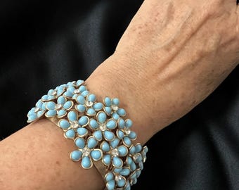 May include: A gold-toned metal cuff bracelet adorned with light blue flower-shaped enamel and clear crystal accents. The bracelet features multiple rows of flowers, creating a bold and elegant statement piece. The bracelet is a fashion accessory.