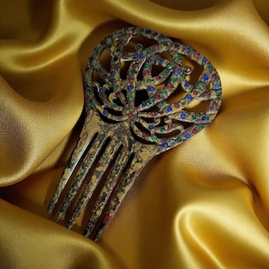 May include: Ornate gold-toned hair comb with a decorative openwork design. The comb is embellished with small, colorful gemstones in red, blue, and green. The comb's tines are long and slender. The background is a golden satin fabric.