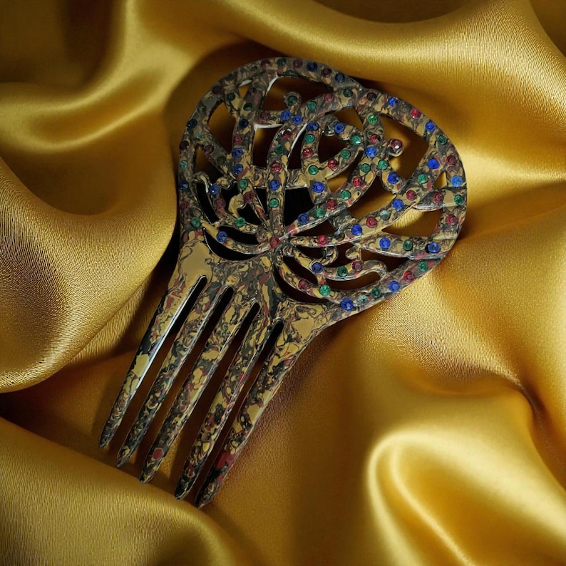 May include: Ornate gold-toned hair comb with a decorative openwork design. The comb is embellished with small, colorful gemstones in red, blue, and green. The comb's tines are long and slender. The background is a golden satin fabric.