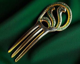 May include: A translucent amber-colored hair comb featuring a decorative openwork top section. The curved upper part is embellished with a series of small, round, vibrant green stones set along the edges and within the swirling cut-out design.