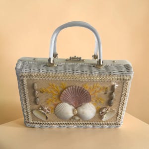May include: A rectangular white wicker handbag featuring a tan woven panel decorated with various seashells and yellow plastic coral-like accents. The bag has a white arched handle, a metallic clasp closure, and gold-toned trim along the edges.