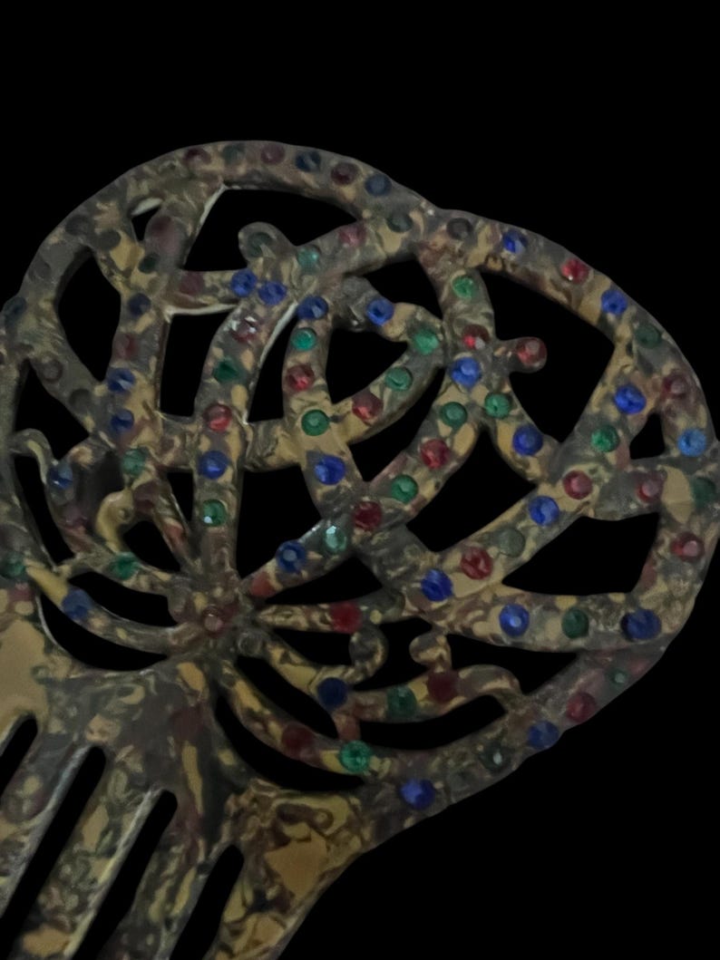 May include: An antique hair comb with a decorative openwork design. The comb is a mottled gold color and is embellished with red, green, and blue rhinestones. The comb's teeth are visible at the bottom of the image.